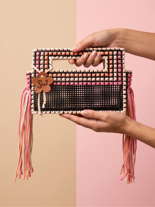 A woven rectangular handbag with a top handle, held by two hands against a split beige and pink background. The bag has a black woven center, pink and beige woven borders, long pink and beige tassels, and a small wooden charm with the word 'Fun'.