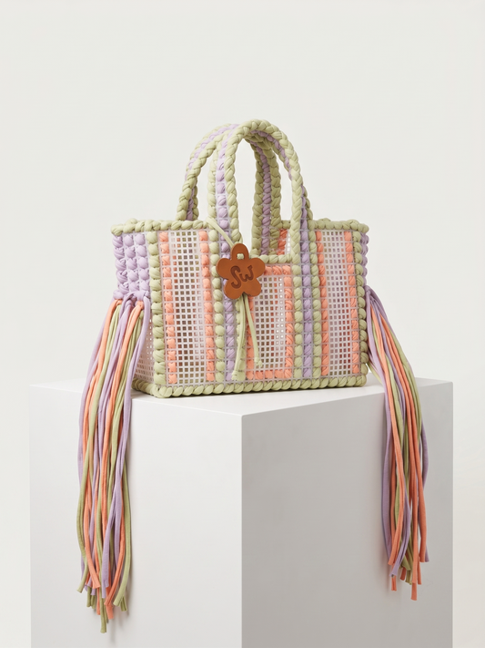 A pastel woven tote bag with braided handles, multicolored fringe tassels, and a small wooden charm, displayed on a white pedestal against a light gray background.