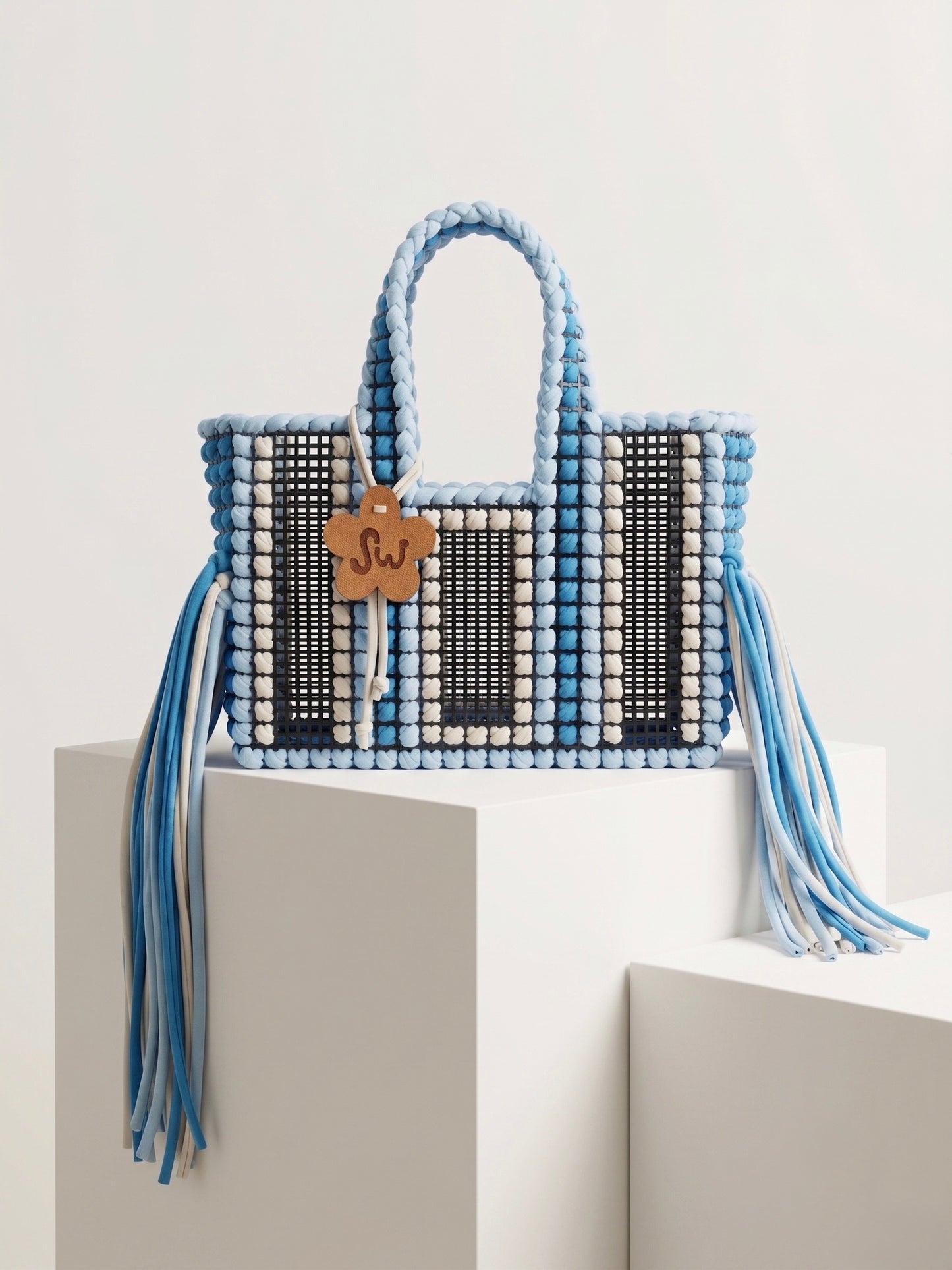 A woven tote bag with light blue and white braided handles and tassels, black and white woven panels, and a small wooden monogram charm reading 'SJ', displayed on white geometric pedestals against a light gray background.