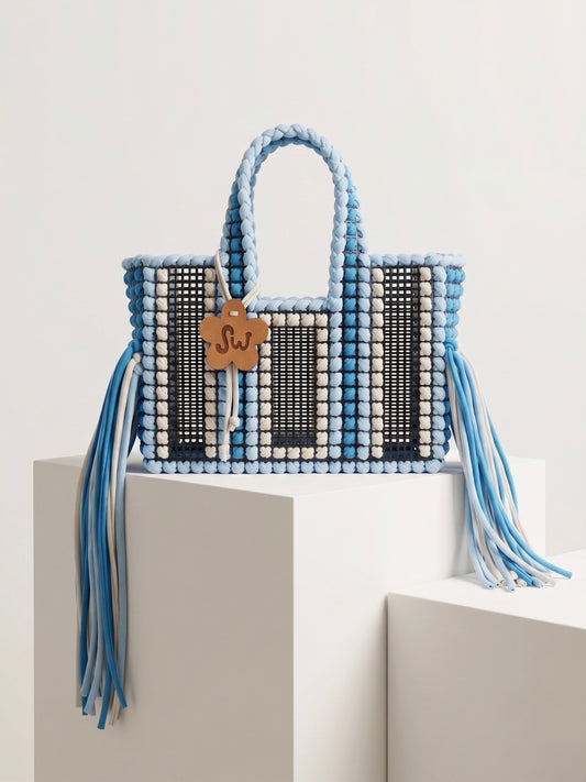 A woven tote bag with light blue and white braided handles and tassels, black and white woven panels, and a small wooden monogram charm reading 'SJ', displayed on white geometric pedestals against a light gray background.